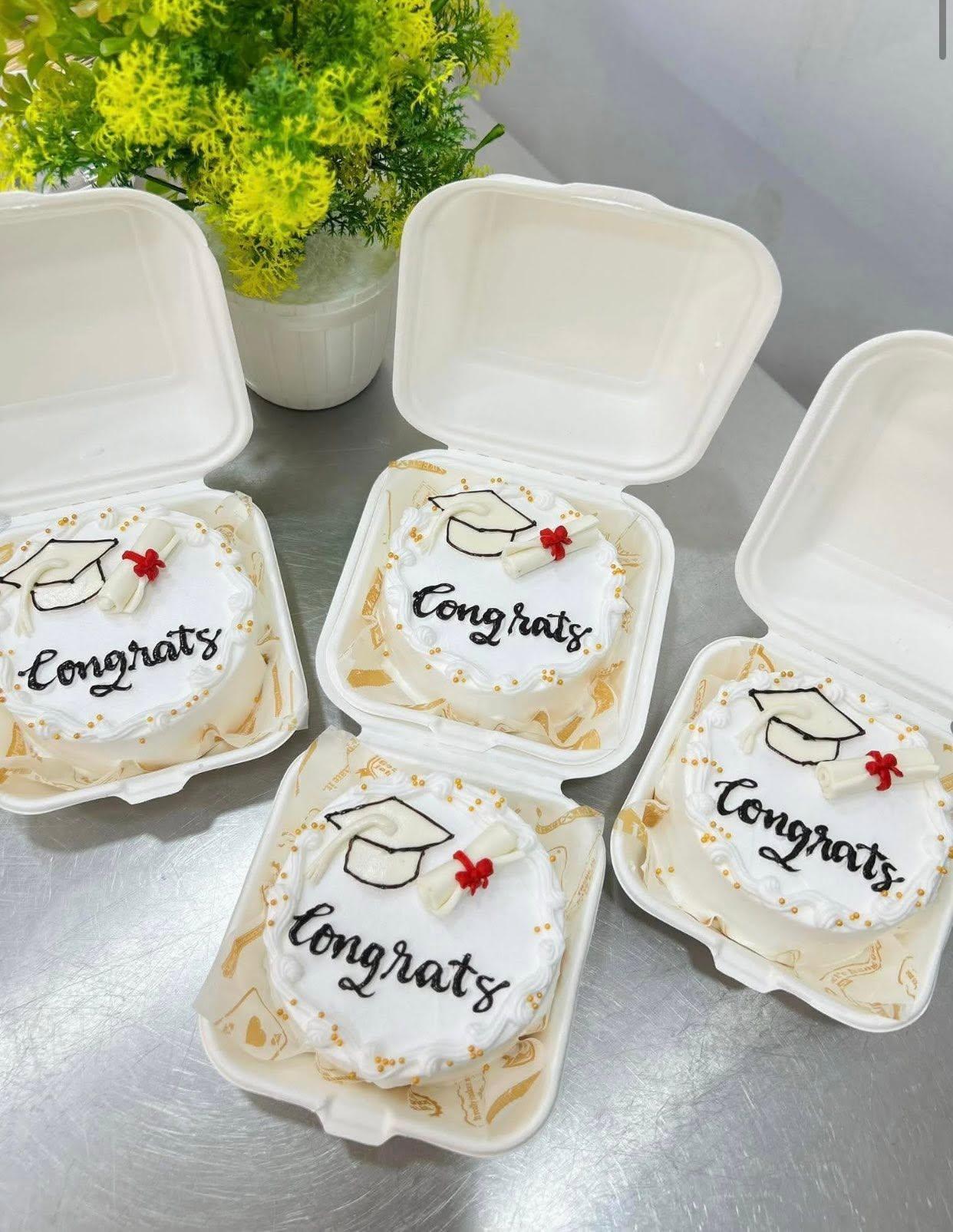 Four small white bento cakes in takeout boxes, each piped with a graduation cap and the word Congrats