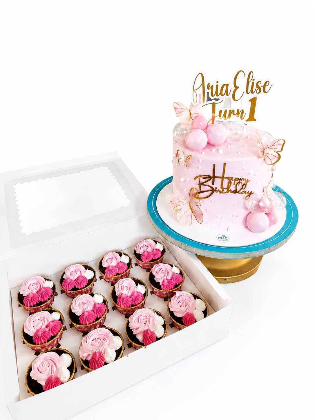 A pink butterfly first-birthday cake with gold script beside an open box of a dozen pink swirl cupcakes