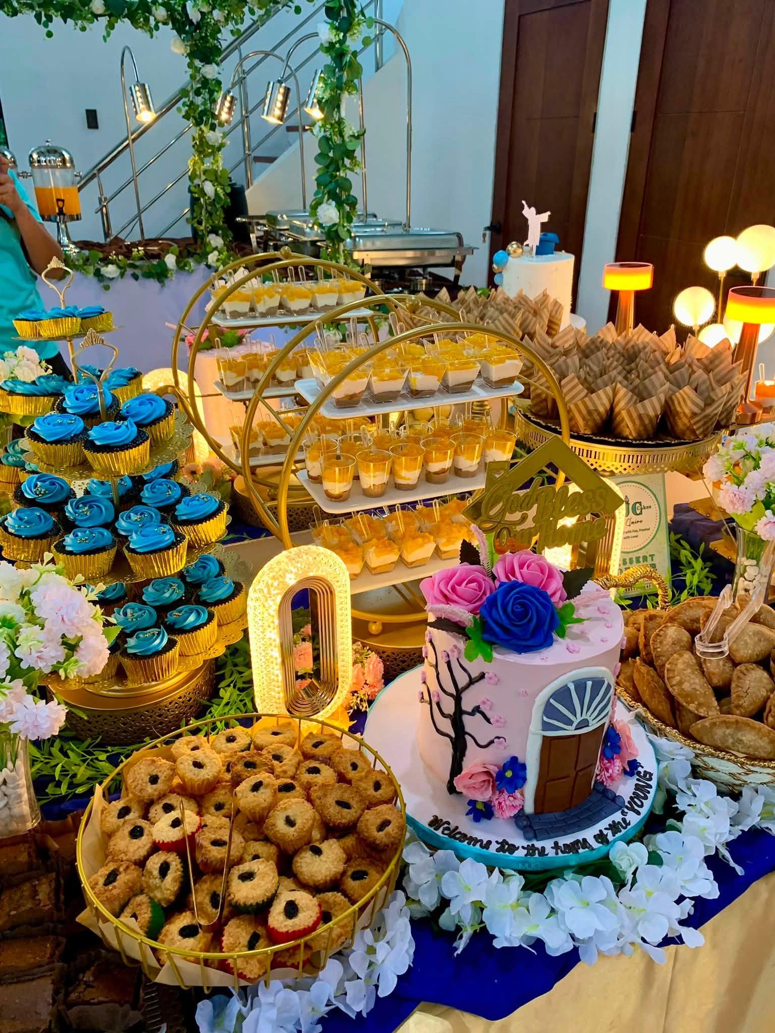 A house blessing dessert table with blue rosette cupcakes, mango float cups, empanada, and a pink house-themed cake