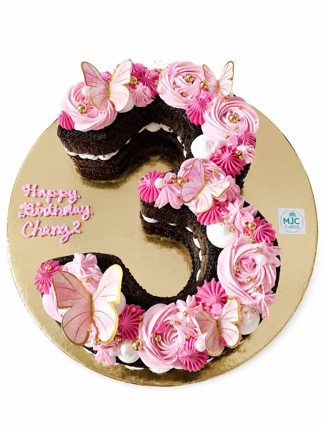 A number three shaped cream cake covered in pink rosettes, pearls, and gold-edged butterflies for Cheng's 3rd birthday
